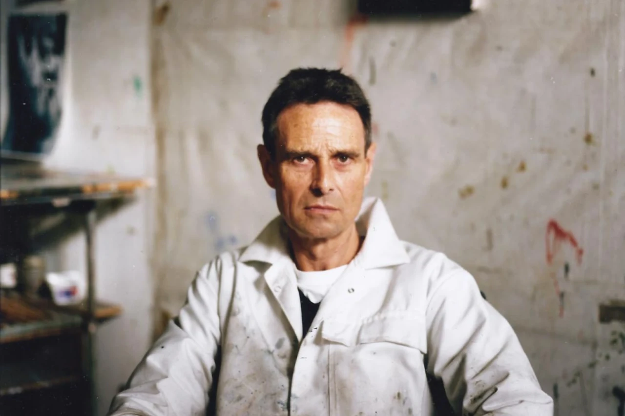 John Keane sat in a chair, wearing a paint spattered white bolier suit,  looking straight at the camera.